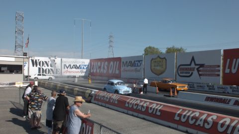 2003 Ford Crown Victoria - small light-colored vintage car and an orange vehicle stage at the start line as spectators watch from the wall, under MavTV and Lucas Oil…