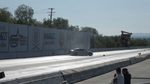 2003 Ford Crown Victoria - silver sports car launches down the drag strip past sponsor wall banners, heading toward the timing scoreboard under power lines.