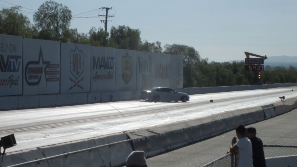 Silver coupe accelerating down the drag strip alongside sponsor banners and a timing scoreboard