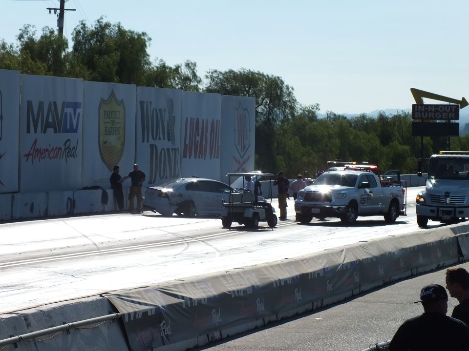 Crashed silver car against the wall with a golf cart, safety truck, and tow at the drag strip