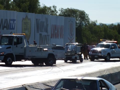 2003 Ford Crown Victoria - flatbed tow truck and golf cart gather at a crash scene against the strip wall, with crew and a safety vehicle attending the damaged car.