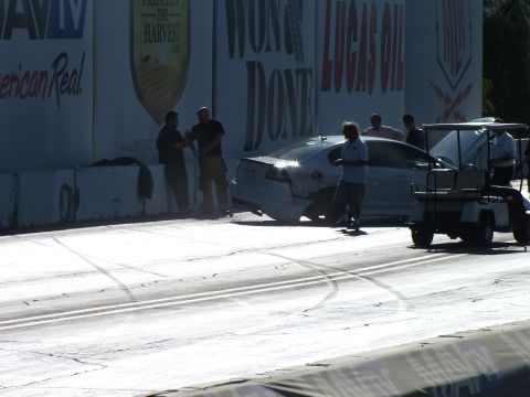 2003 Ford Crown Victoria - damaged silver car sits against the wall as crew members and a golf cart attend it, with dark tire marks streaking the drag-strip surface.