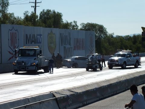 2003 Ford Crown Victoria - wide view of the crash recovery shows a tow truck, golf cart, and safety pickup clustered around a damaged car at the drag-strip wall.