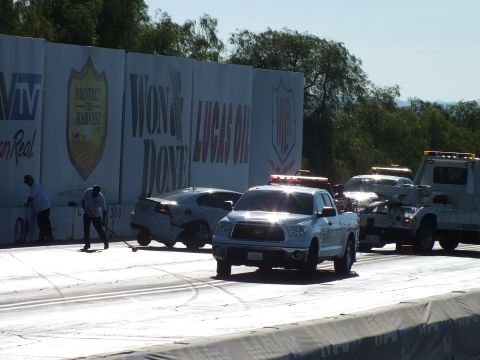 2003 Ford Crown Victoria - white Toyota pickup and a tow truck work to remove a damaged car wedged against the drag-strip wall while a worker sweeps debris.