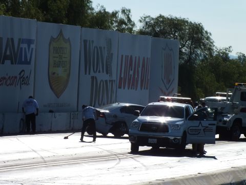2003 Ford Crown Victoria - worker sweeps debris while a white Toyota pickup and tow truck clear a wrecked car from the drag-strip wall under sponsor banners.