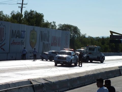 2003 Ford Crown Victoria - white pickup and a flatbed tow truck stop on a sunlit drag strip lined with Lucas Oil and MAVTV banner walls.
