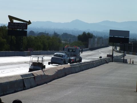 2003 Ford Crown Victoria - tow truck pulls a small car along a drag strip lane with hazy mountains and an In-N-Out sign in the background.