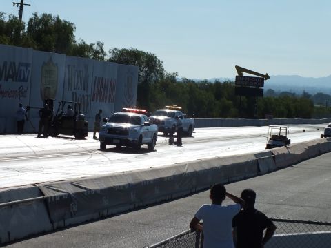 2003 Ford Crown Victoria - White trucks sit on a bright drag strip beside a banner wall while spectators watch from behind a chain-link fence.