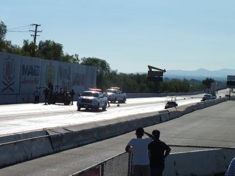 2003 Ford Crown Victoria - Wide view of a drag strip with white trucks at the start area and spectators standing along the fence in the foreground.