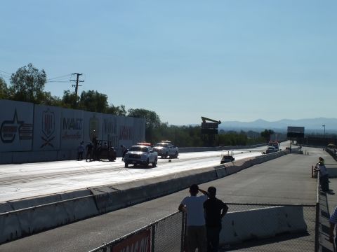 2003 Ford Crown Victoria - long drag strip stretches toward hazy hills with white trucks at the lights and spectators watching from the rail.