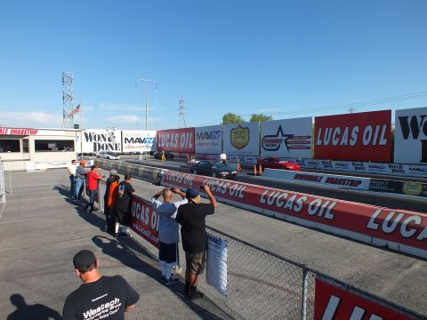 2003 Ford Crown Victoria - crowd gathers at the rail of Irwindale Dragstrip watching staged cars, with power lines and sponsor walls behind.