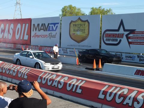 2003 Ford Crown Victoria - white sedan and a black Dodge Charger sit staged side by side on the drag strip in front of Protect the Harvest banners.