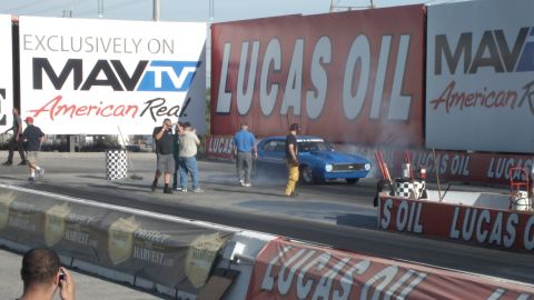 2003 Ford Crown Victoria - blue muscle car smokes its tires near the start line as photographers and crew watch under a MAVTV banner.