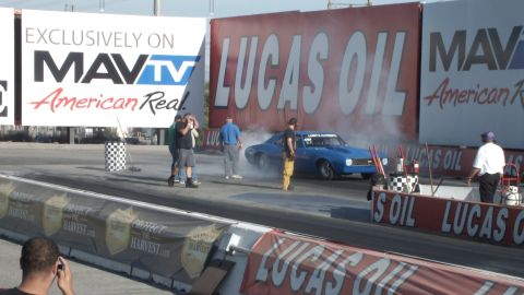 2003 Ford Crown Victoria - blue muscle car spins up in tire smoke at the start line as crew and photographers gather along the Lucas Oil banner.