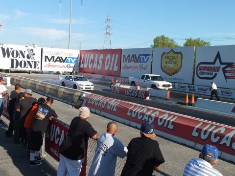 2003 Ford Crown Victoria - white sedan and a white pickup launch down the drag strip lanes as spectators watch from the fence under sponsor banners.