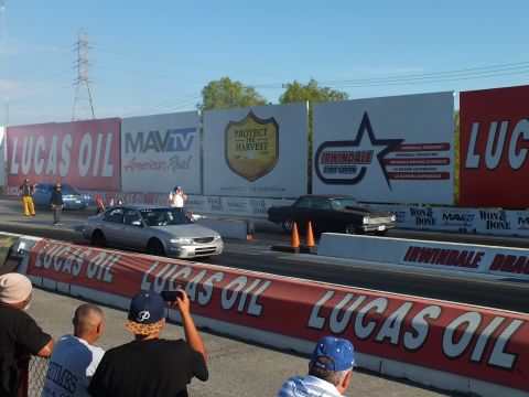 2003 Ford Crown Victoria - silver sedan and a dark muscle car race down the drag strip lanes as a crowd watches from behind the Lucas Oil rail.