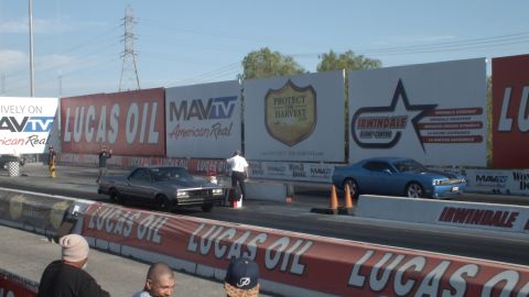 2003 Ford Crown Victoria - dark sedan and a blue Dodge Challenger sit staged on the drag strip in front of Irwindale and Lucas Oil banners.