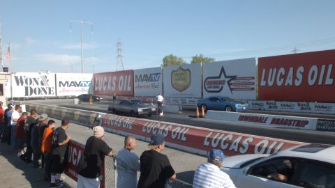 2003 Ford Crown Victoria - dark car launches down the drag strip past a crowd lining the fence under Won & Done and Lucas Oil banners.