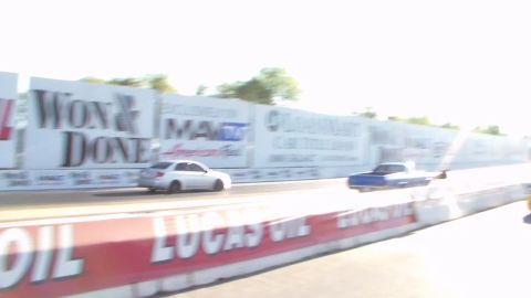 2003 Ford Crown Victoria - silver sedan and a blue car race down a sunlit drag strip past sponsor banners and Lucas Oil barriers.