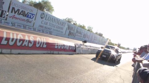 2003 Ford Crown Victoria - dark car accelerates down a drag strip lined with sponsor banners as spectators watch from the rail.