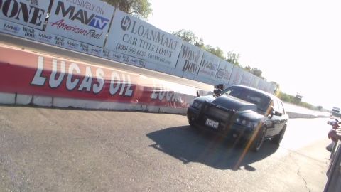 2003 Ford Crown Victoria - black sedan launches down a drag strip beside a Lucas Oil barrier wall and sponsor banners.