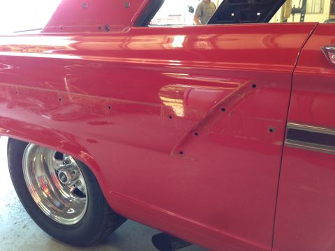 1964 Ford Fairlane - Close-up of a red vintage car's side, showing drilled holes and chrome trim, with a person visible in the background.
