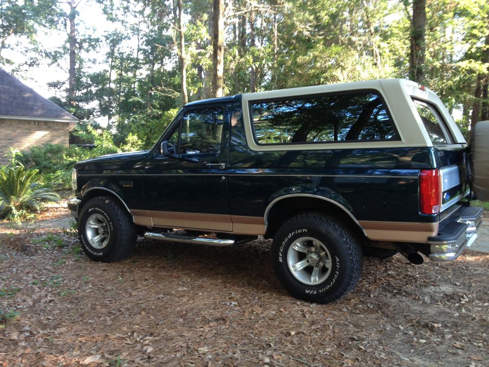 Dark green and tan Ford Bronco SUV, side view, parked outdoors on a dirt and leaf-covered surface