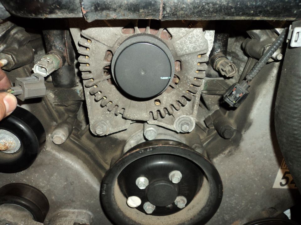Close-up of a car's alternator with a pulley below it. A hand is disconnecting a wire harness