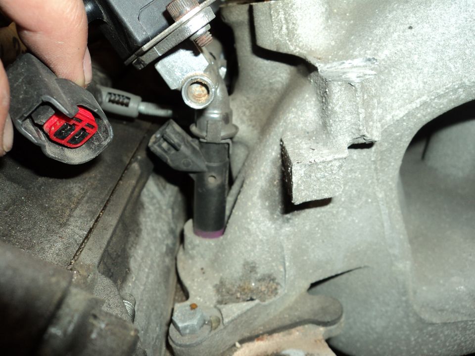 Hand holding red-tabbed electrical connector near engine component with purple-tipped part