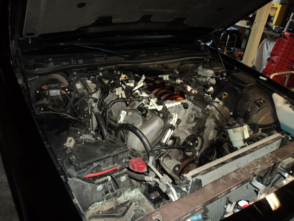Car engine bay with hood open, showing the engine and various hoses and wires. Some parts are disconnected