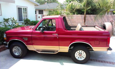 1994 Ford Bronco - Driver's-side profile of a red and tan two-tone full-size Ford Bronco 4x4 with the removable hardtop taken off, exposing the tan interior,…