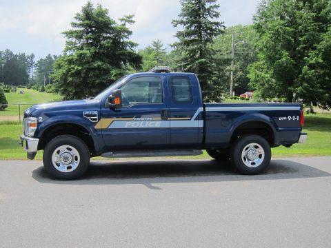2001 Ford Crown Victoria - A blue police pickup truck is parked on an asphalt surface with trees and a fence in the background.