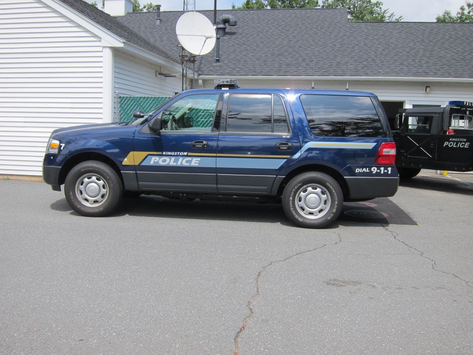 Blue Ford Expedition police SUV with 'KINGSTON POLICE' markings and 'DIAL 9-1-1' on rear quarter panel. Another black police vehicle is partially visible to the right