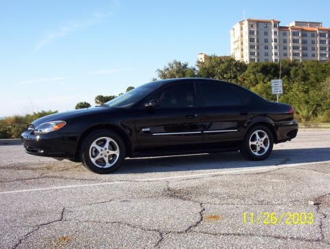 2003 Ford Taurus - A black 2003 Ford Taurus sedan is parked on a cracked asphalt surface with a building and trees in the background.