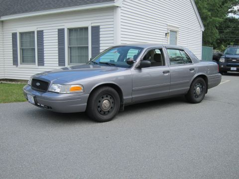 2001 Ford Crown Victoria - A gray 2001 Ford Crown Victoria sedan, likely a police vehicle, is parked in front of a white building.