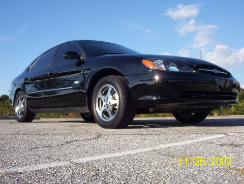 2003 Ford Taurus - A black Ford Taurus sedan photographed from a low front three-quarter angle on a paved lot under a blue sky with scattered clouds. It has a…