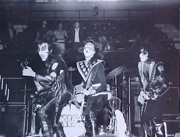 Band members in costume and makeup play instruments on stage in front of a drum kit with 'KISS' logo