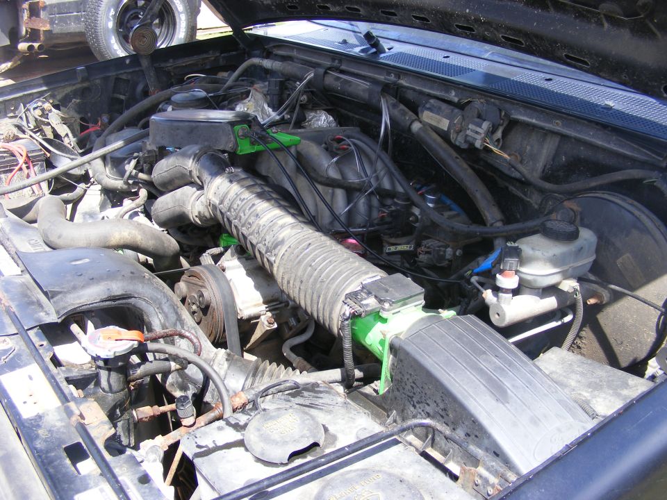 Engine bay with hoses, belts, and air intake system visible. Another vehicle's wheel is in the background