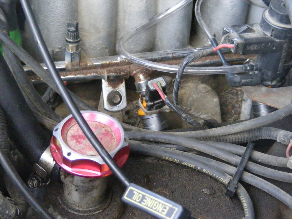 Close-up of a vehicle's engine bay, showing the fuel rail, injectors, and various hoses. An aftermarket red oil cap is visible