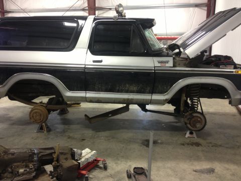 1979 Ford Bronco - A two-tone Ford Bronco is raised on jack stands in a garage, with its hood open, revealing the engine bay.