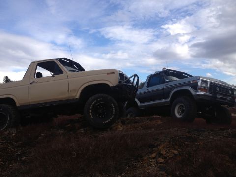 1979 Ford Bronco - Two modified Ford Broncos, likely from the late 1970s, are positioned on uneven terrain under a partly cloudy sky.