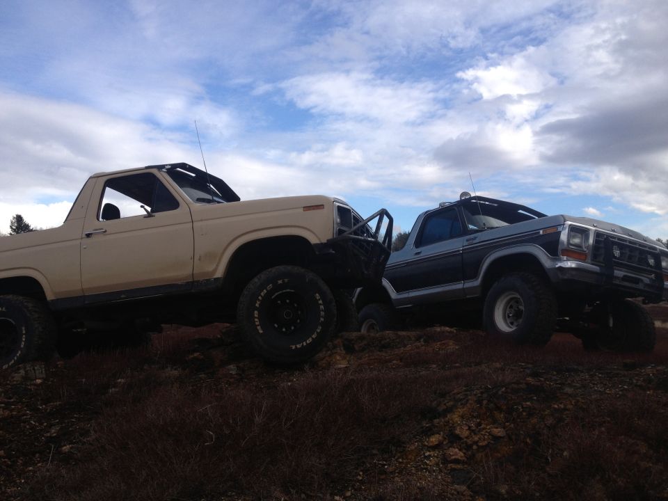 Tan 1978 Ford Bronco and a blue 1978 Ford Bronco on a dirt hill under a cloudy sky