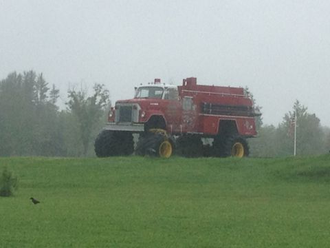 1979 Ford Bronco - Modified fire truck with massive tires and yellow wheels sits on a green hill under a cloudy sky.