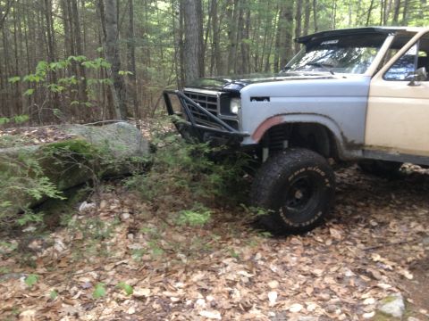 1979 Ford Bronco - This modified pickup truck is parked off-road in a wooded area, next to a large rock.
