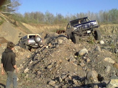 1979 Ford Bronco - Two modified SUVs are shown climbing a challenging, rocky incline under a clear blue sky.