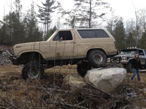 1979 Ford Bronco - A tan 1979 Ford Bronco is shown off-roading, climbing over a large rock with another SUV visible in the background.