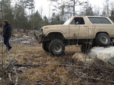 1979 Ford Bronco - A person walks past a lifted, tan SUV that is positioned on large rocks in a wooded area.