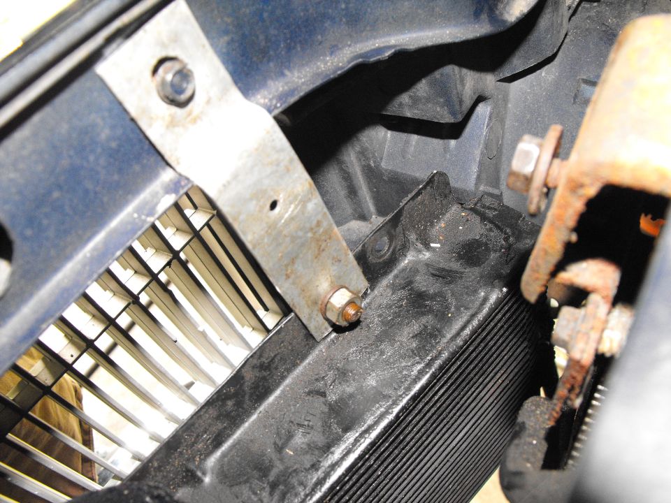 Close-up of a metal bracket bolted behind a car's grille and core support, with rusty hardware nearby