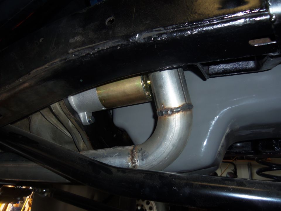 Close-up of a vehicle's undercarriage showing a starter motor connected to an exhaust pipe. Welds are visible on the pipe