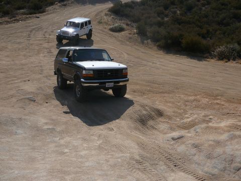 1988 Ford Bronco - Two off-road vehicles, a Ford Bronco and a white Jeep, navigate a dusty trail in a dry, hilly landscape.
